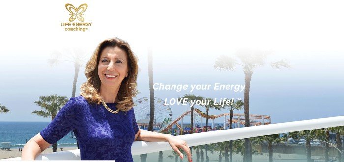 这张Life Energy Coaching主页的截图上面有一个金色的标志，上面是一张穿着蓝色衬衫的黑发女人微笑着倚靠在俯瞰海滨游乐园的阳台上的照片，旁边还有白色的文字，邀请顾客改变他们的能量，热爱他们的生活。