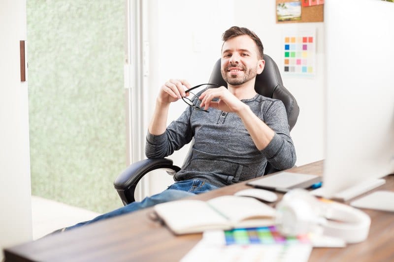 This photo shows a smiling man sitting in an office chair near a desk with color tables, a computer monitor, and other graphic design tools.