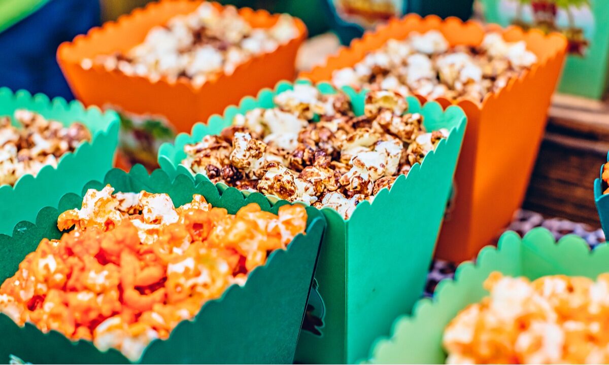 Buckets of different flavors of gourmet popcorn sit side by side in orange and green boxes.