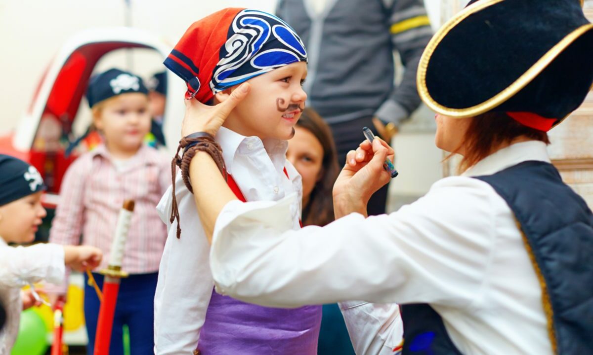 A woman in a pirate outfit paints a mustache on a child attending a party.