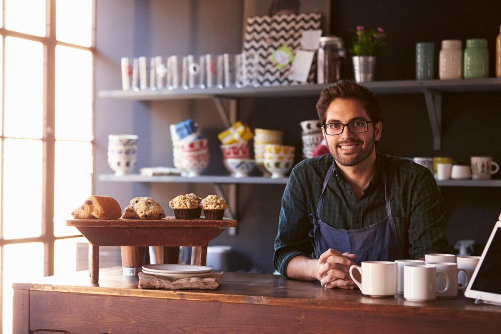 年轻的企业家坐在自己的咖啡店里微笑着，背后是咖啡杯