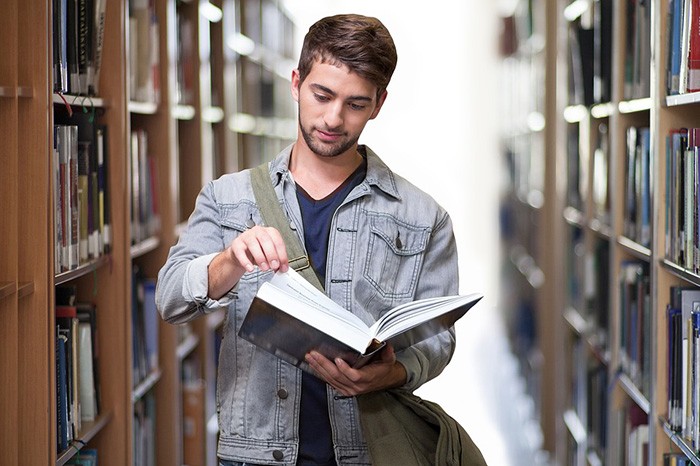 Younger man reading a book in a college library as an example of an undergraduate student