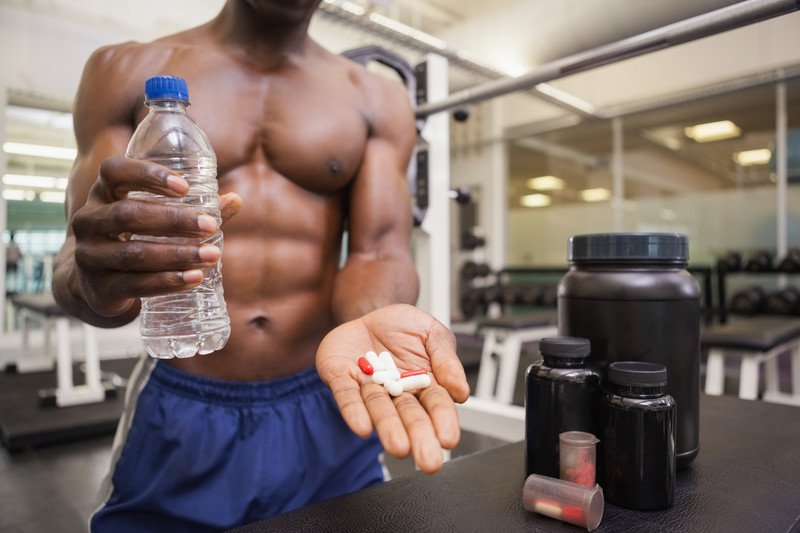 This photo shows a shirtless man in blue shorts inside a gym, holding a bottle of water in one hand and a handful of pills in the other, representing the best sports supplements affiliate programs.