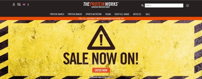 This screenshot of the home page for The Protein Works shows an image of what looks like a black and yellow hazard sign, with text in black lettering that reads 'Sale Now On!'