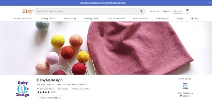 这张Baby Q设计主页的截图显示了一顶玫瑰色的无边帽，上面有几个绿色、橙色、红色、黄色和粉色的毛毡球，下面是一个白色的标题，上面有Etsy的标志和一个蓝色的栏，宣布在Etsy上出售的许多商品都可以免费送货。