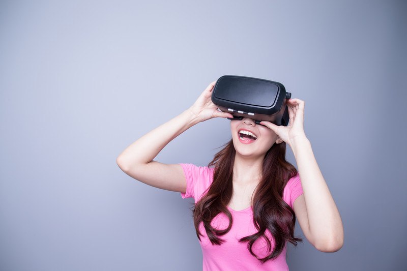 这张照片是一个微笑的女人，棕色的长发，粉红色的衬衫，拿着一套虚拟现实眼镜到她的眼睛，在灰色的背景前，代表了最好的虚拟现实联盟计划。