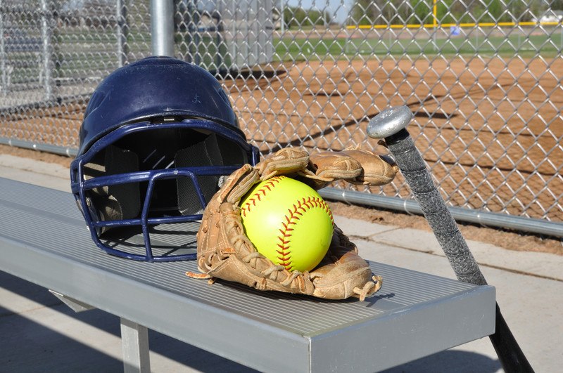 这张照片展示了一个深蓝色的头盔，一个棕色的手套，一个黑色的球棒，和一个黄色的垒球在铝制板凳上或附近的一个链环围栏附近的垒球场，代表最好的垒球联盟计划。