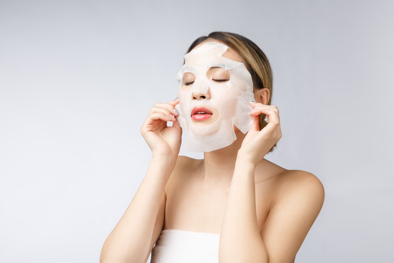 This photo shows a young Asian woman in a white towel, standing in front of a white background, applying a white sheet mask, representing the best Korean beauty products affiliate programs.
