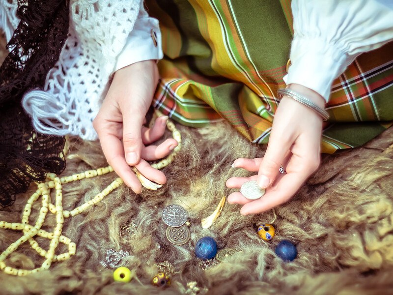 这张照片显示了一个年轻女子的手，她正在整理各种珠子、硬币和符文，仿佛准备施咒，代表着最好的巫术附属项目。