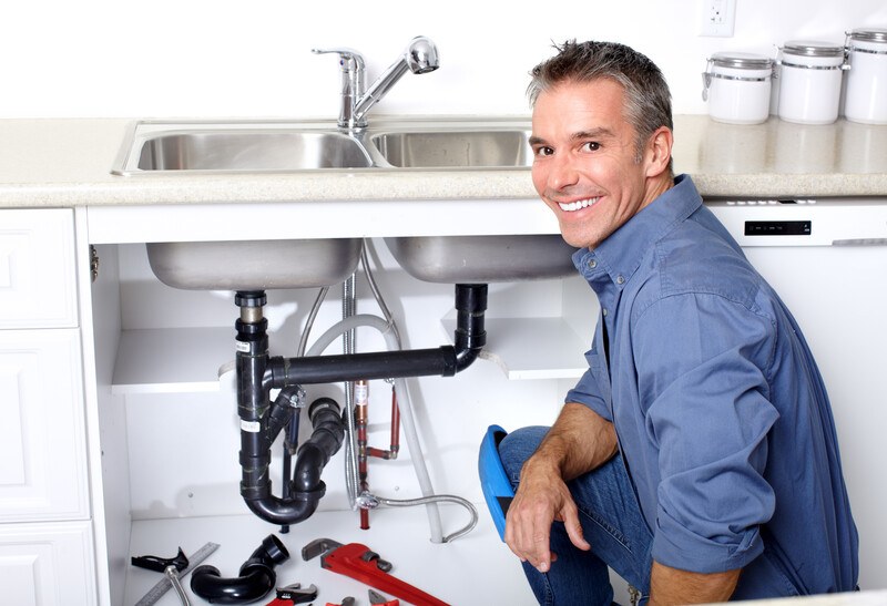 This photo shows a smiling man in a blue shirt and jeans kneeling next to an open sink and tools, representing the question, do plumbers make good money?