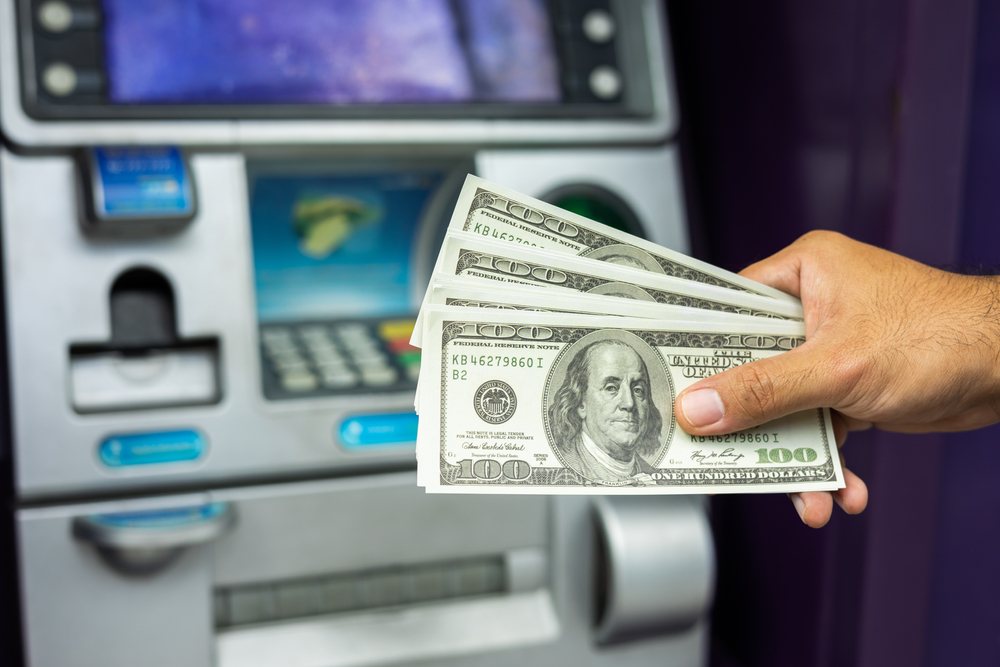 一个米an's hand holds a wad of hundred dollar bills in front of a freestanding ATM.