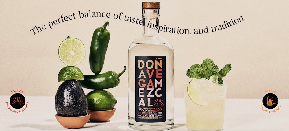 A bottle of Dona Agave Mezcal sits next to a margarita on one side and limes and jalapenos on the other.