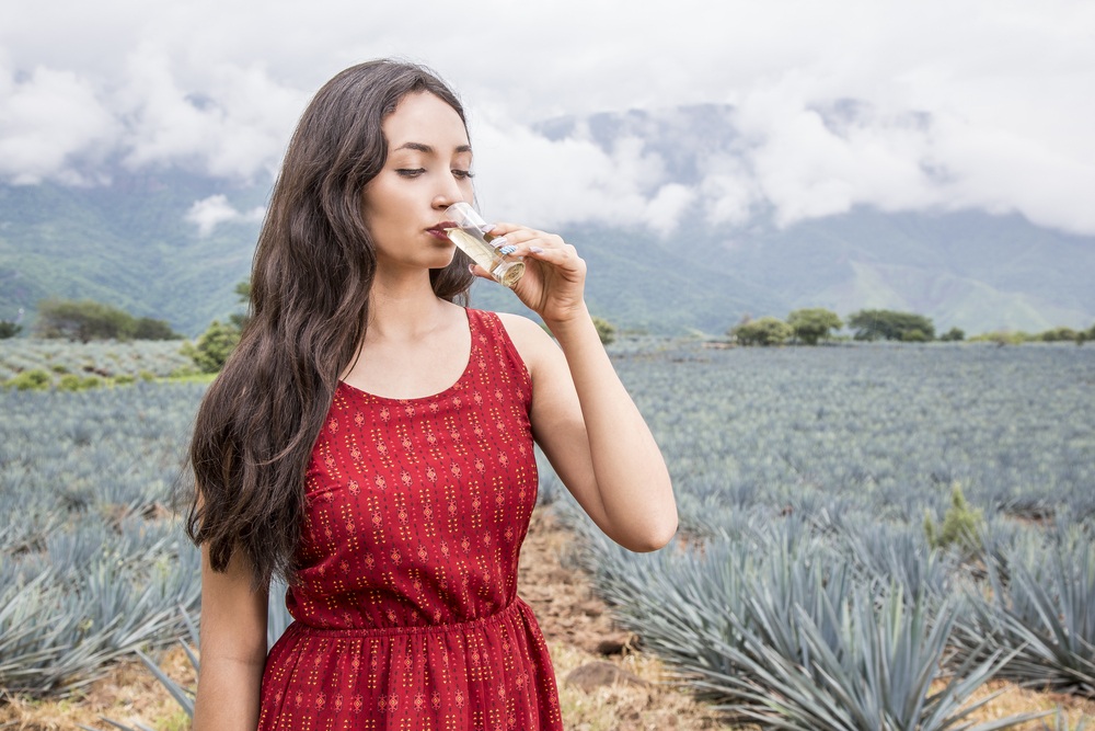 woman drinks a glass of tequila in an agave field.