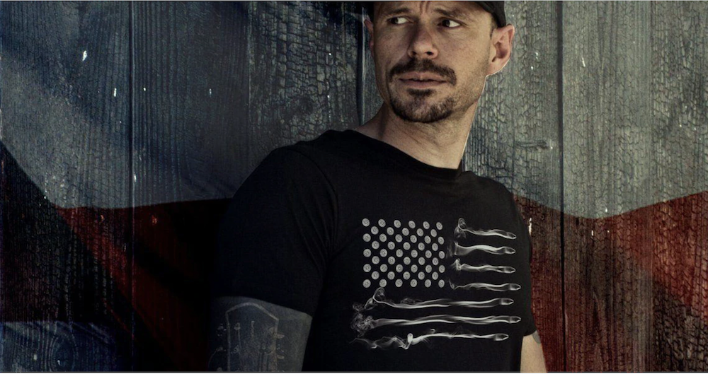 A man in a black American flag t-shirt poses against a red and gray wooden wall.