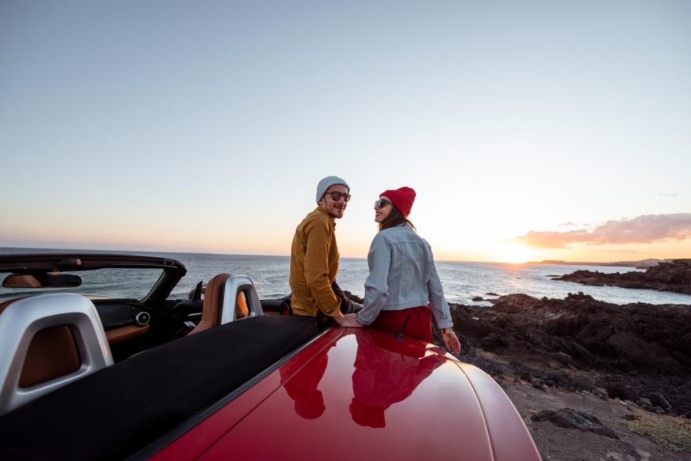 年轻的男人和女人坐在g on the side of a car looking over the ocean